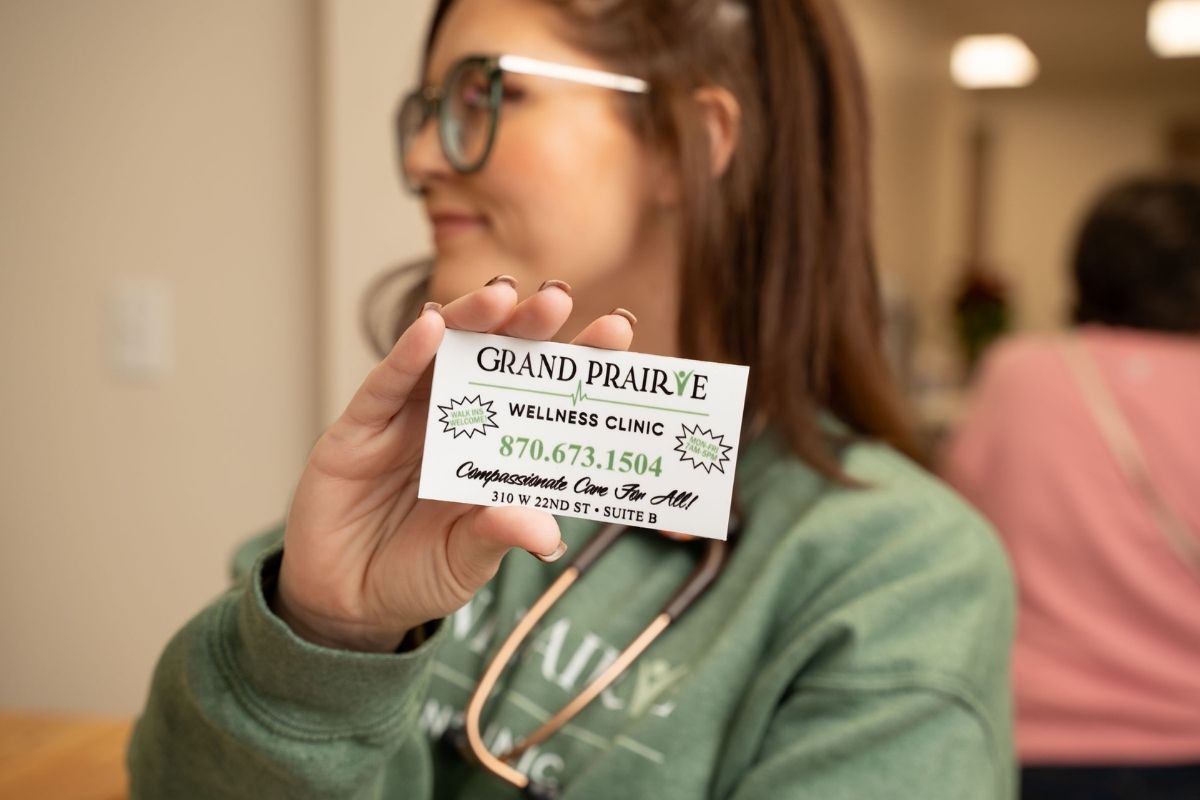 A woman wearing glasses and a stethoscope holds up a business card for Grand Prairie Wellness Clinic, displaying the phone number and address, with blurred people in the background.
