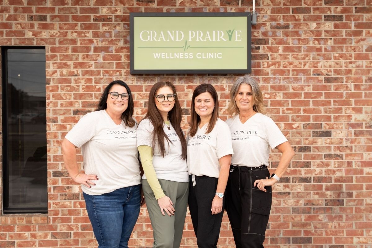 Four women wearing matching Grand Prairie Wellness Clinic shirts stand smiling in front of a brick building under a sign reading Grand Prairie Wellness Clinic.