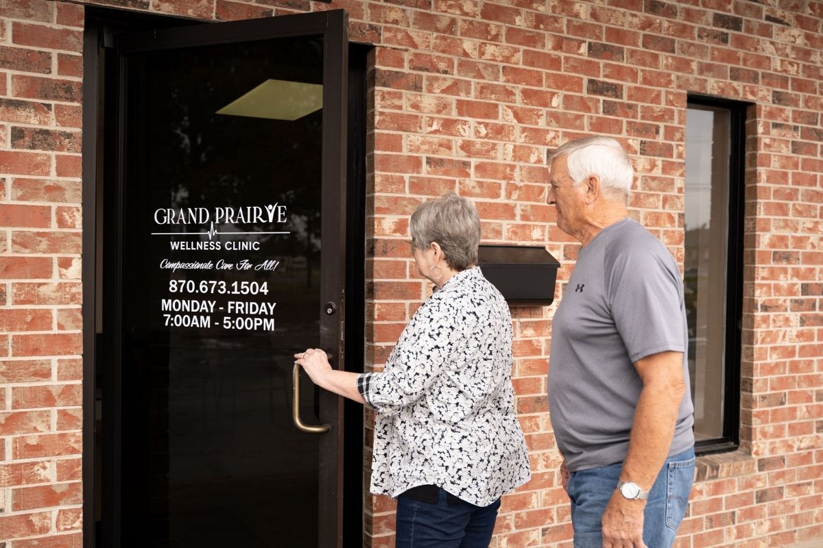 An older woman and man stand outside a brick building. The woman is opening a glass door labeled Grand Prairie Wellness Clinic, displaying the clinics hours, phone number, and slogan.