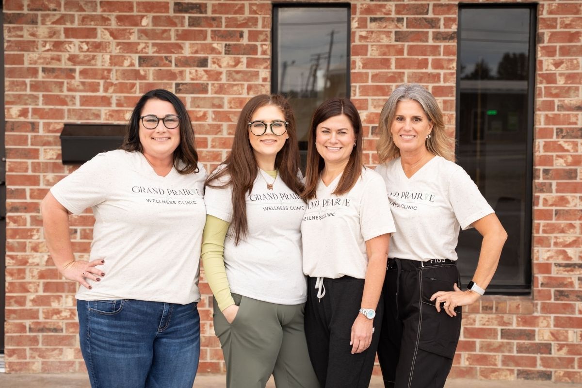 Four women stand side by side smiling in front of a brick wall, all wearing matching light-colored Grand Prairie Wellness Clinic T-shirts and casual pants.