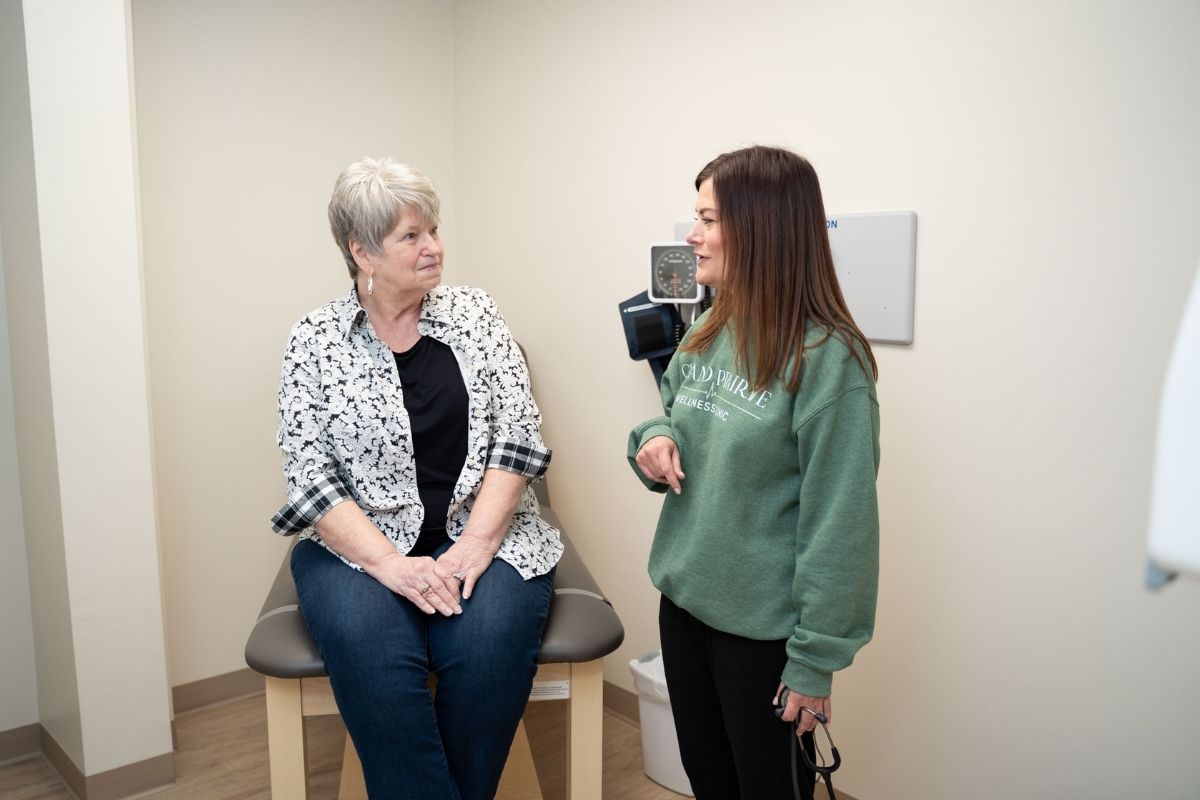 An older woman sits on an exam table in a medical office, talking to a younger woman standing nearby. Both appear engaged in conversation. The room has neutral-colored walls and medical equipment is visible.