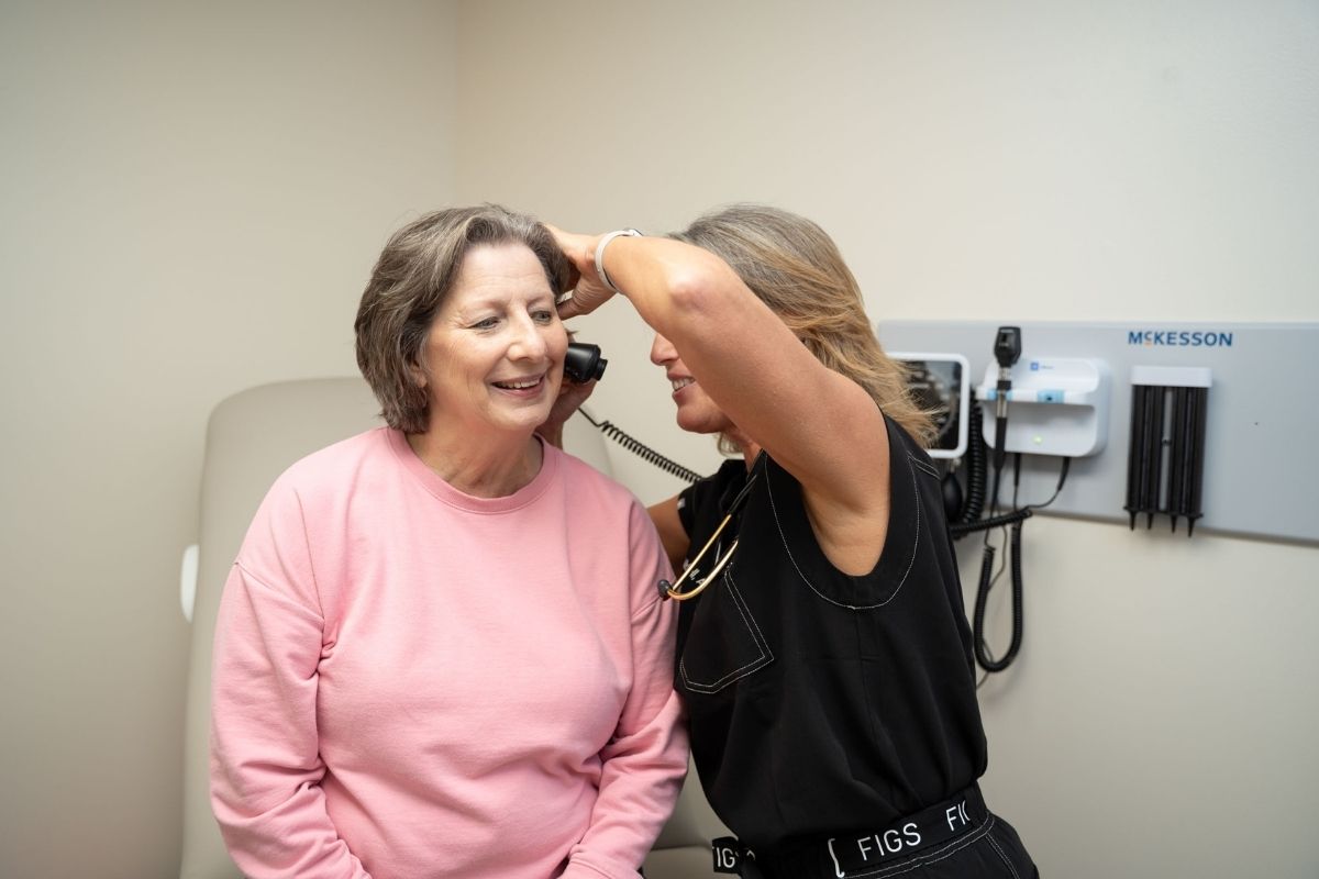 A healthcare professional examines an older woman’s ear with an otoscope in a medical office. The woman is seated and smiling, wearing a pink sweater. Medical equipment is visible on the wall behind them.