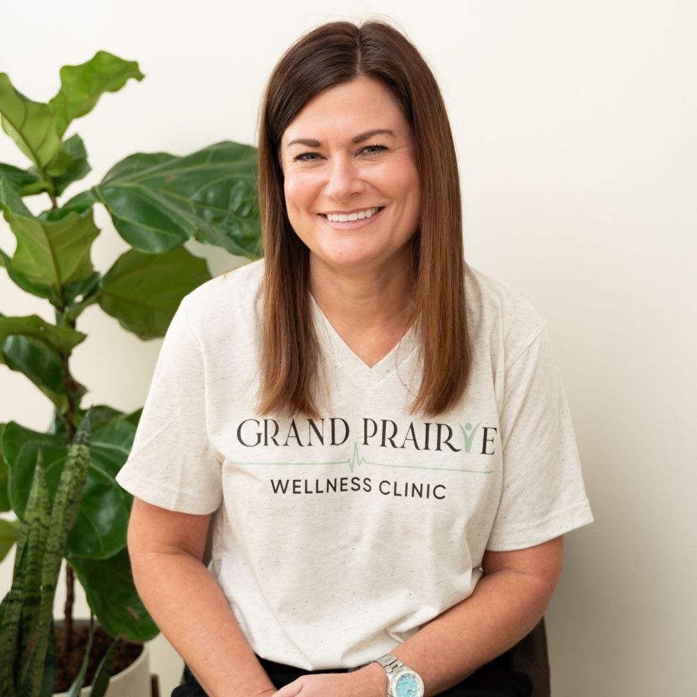 A woman with straight brown hair, wearing a Grand Prairie Wellness Clinic T-shirt, sits and smiles in front of a light-colored wall with green leafy plants beside her.