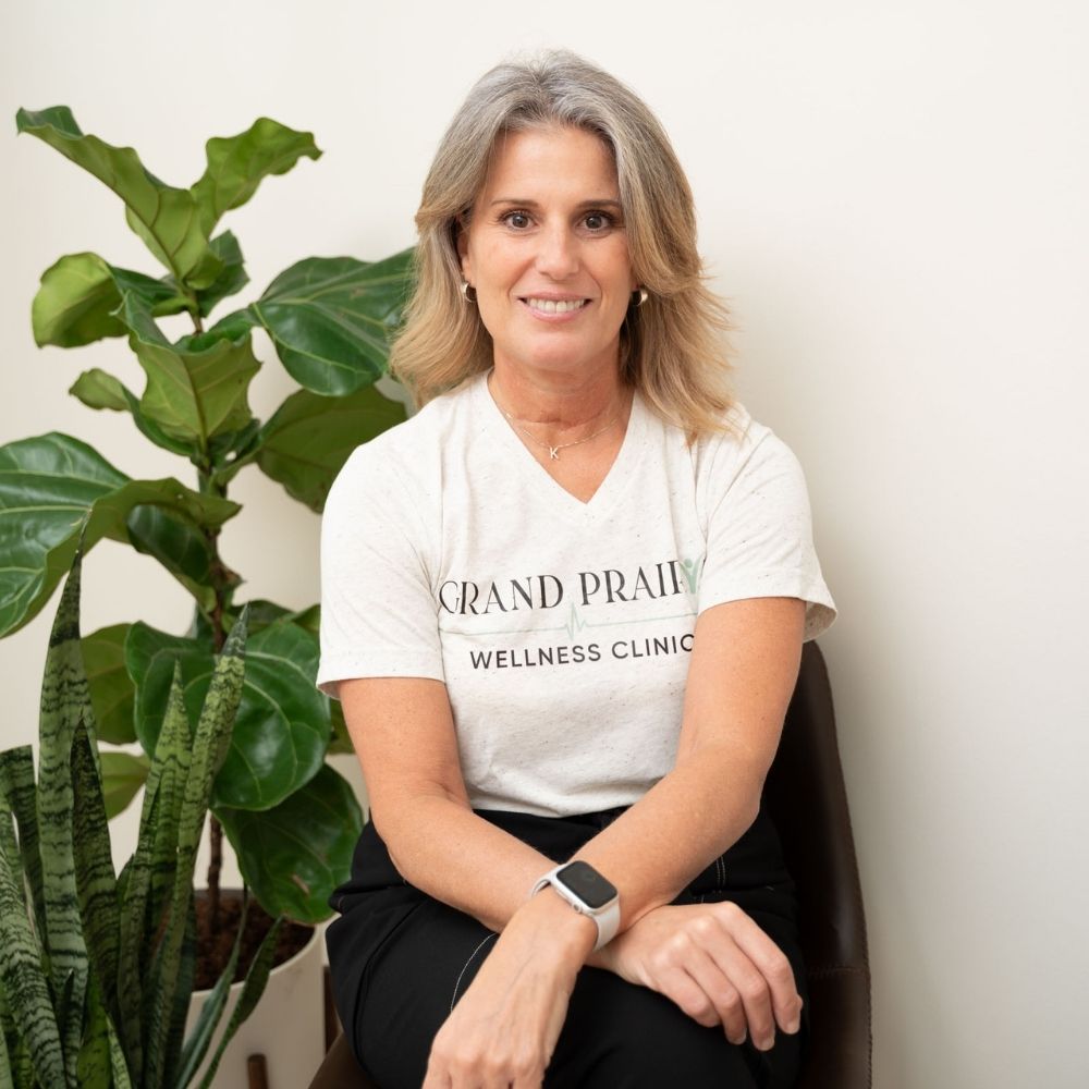 A woman with blonde hair sits on a brown chair, smiling. She wears a Grand Prairie Wellness Clinic shirt, black pants, and a smartwatch. Two green plants are in the background against a plain, light-colored wall.
