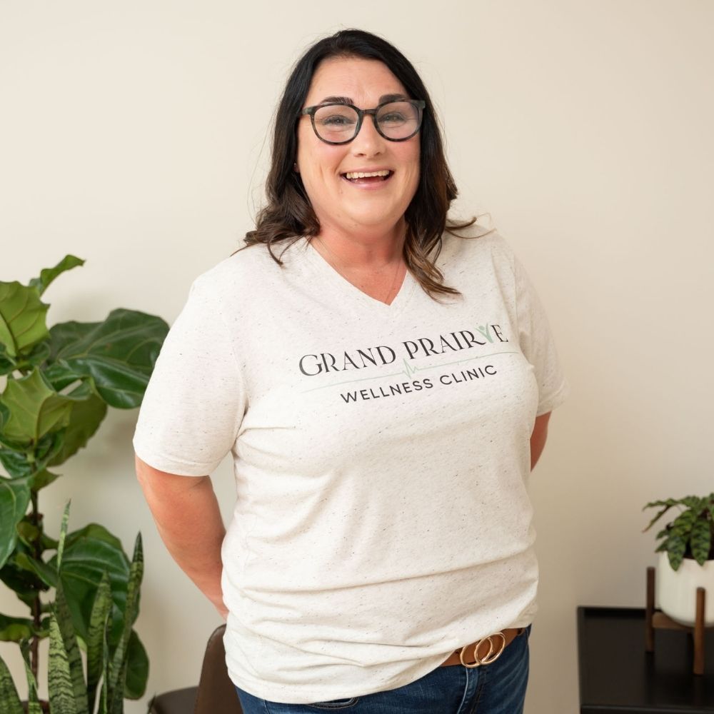 A woman with long dark hair and glasses smiles while standing indoors. She is wearing a white Grand Prairie Wellness Clinic t-shirt. Green potted plants are visible in the background.