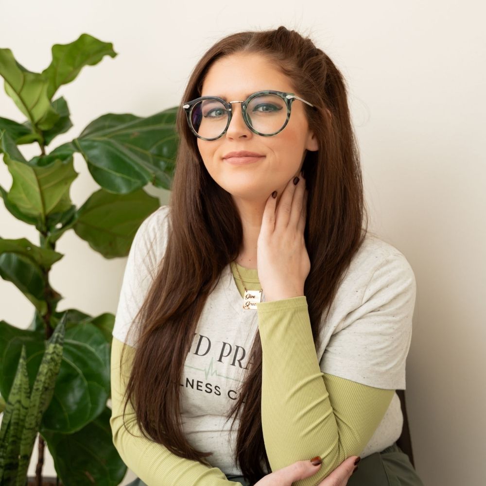 Woman with long brown hair and glasses sits in front of a green leafy plant, wearing a light-colored shirt and green long sleeves underneath, gently touching her neck and smiling softly at the camera.