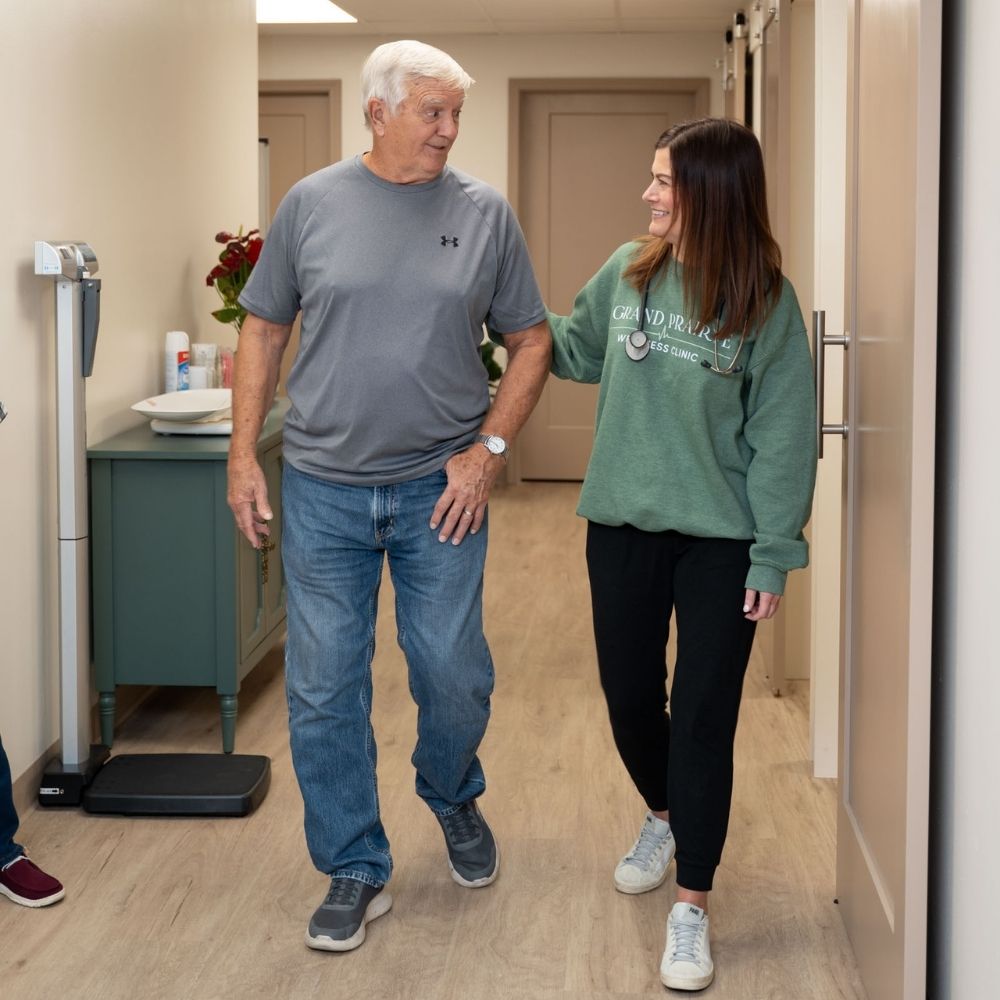 An older man in a gray shirt and jeans walks down a hallway, smiling and talking with a younger woman in a green sweatshirt and black pants. They appear to be in a medical or wellness facility.