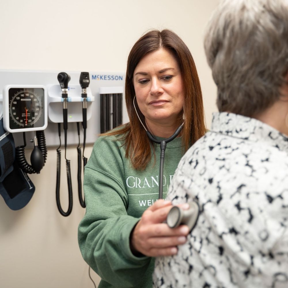 A healthcare professional listens to an older patients back with a stethoscope in a medical examination room. Medical instruments are mounted on the wall in the background.