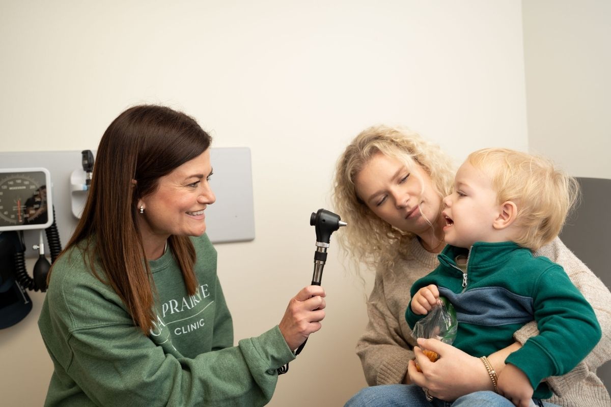 A female healthcare provider smiles while holding an otoscope and interacting with a young boy sitting on a womans lap during a medical exam in a clinic room.