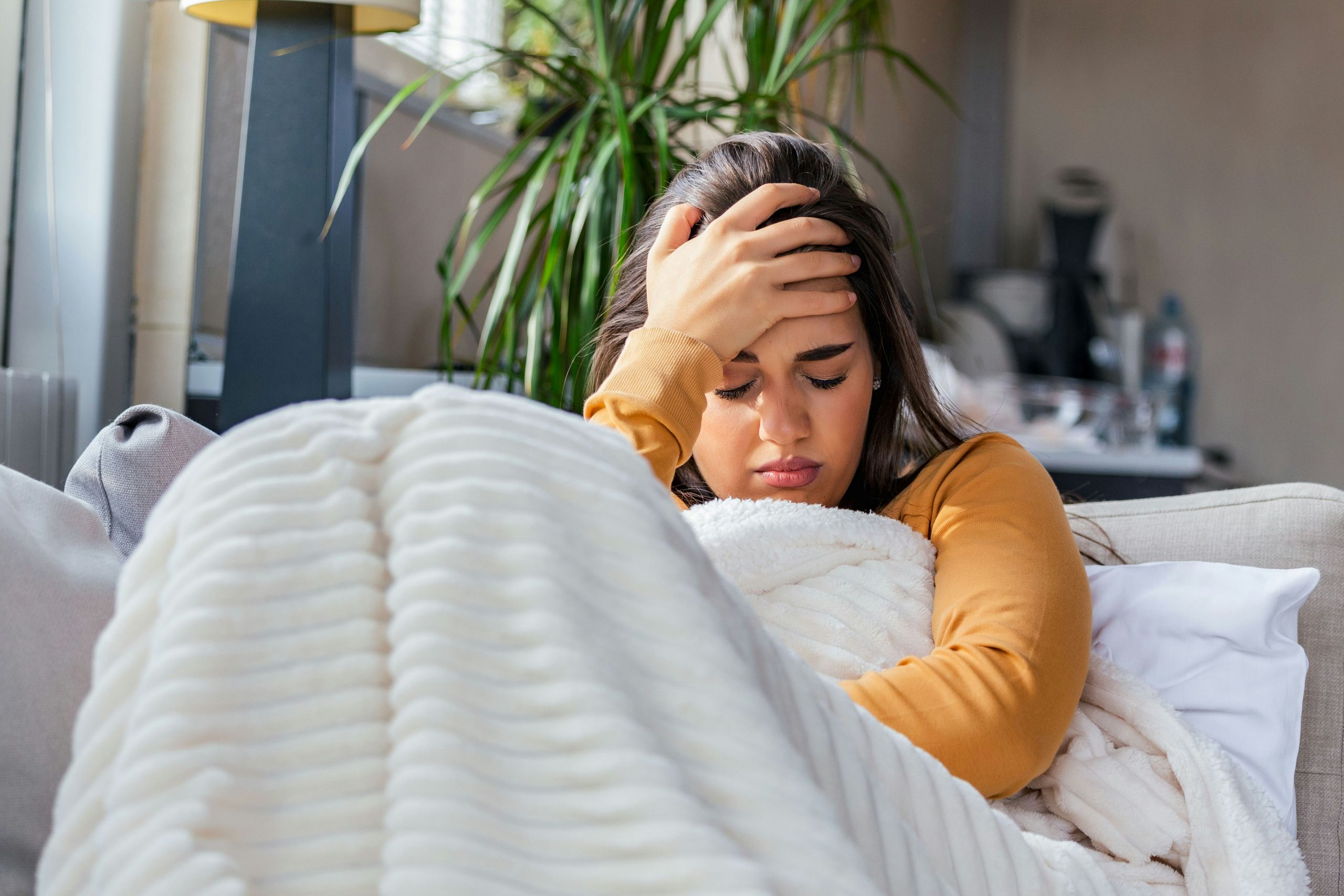 A woman in a mustard yellow sweater sitting on a couch, holding her head with a pained expression while wrapped in a white blanket.